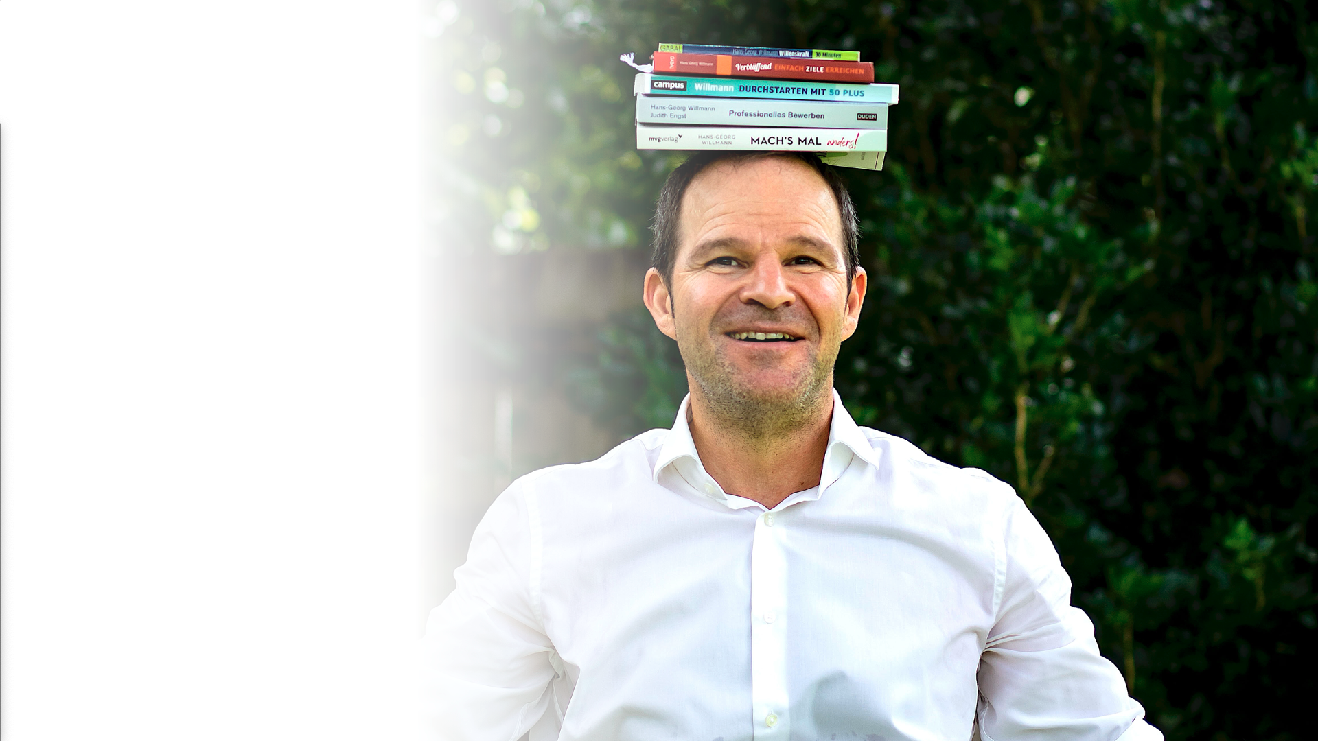 Ein Mann in einem weißen Hemd steht im Freien, lächelnd, mit einem Stapel Bücher auf dem Kopf. Im Hintergrund ist grünes Laub zu sehen.