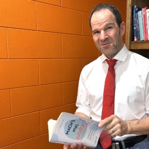 Ein Mann mit weißem Hemd und roter Krawatte hält ein aufgeschlagenes Buch mit dem Titel Machs mal anders! neben einem hölzernen Bücherregal vor einer orangefarbenen Backsteinwand.