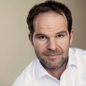 A middle-aged man with short dark hair and a beard wears a white shirt and looks directly at the camera against a plain background.